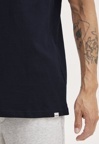 Tricou din bumbac bleumarin, cu un design simplu, mâneci scurte și o etichetă albă mică cusută la tiv. Materialul gri al pantalonilor este vizibil.