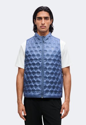 Homme portant un gilet matelassé sans manches bleu par-dessus une chemise blanche à manches courtes, debout devant un fond clair uni.