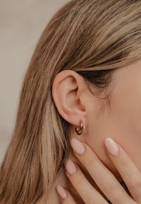 Ohrringe aus Roségold in Form von Ohrringen mit einer glatten, glänzenden Oberfläche und einer kleinen Reihe von klaren Edelsteinen. Die Hand des Models berührt sanft ihren Hals.