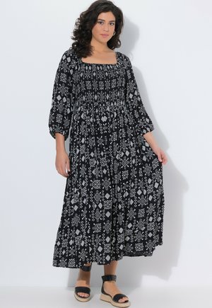 Kobieta ubrana w czarną i białą sukienkę z wzorem do kostek, z rękawami do łokcia i czarne sandały na platformie.