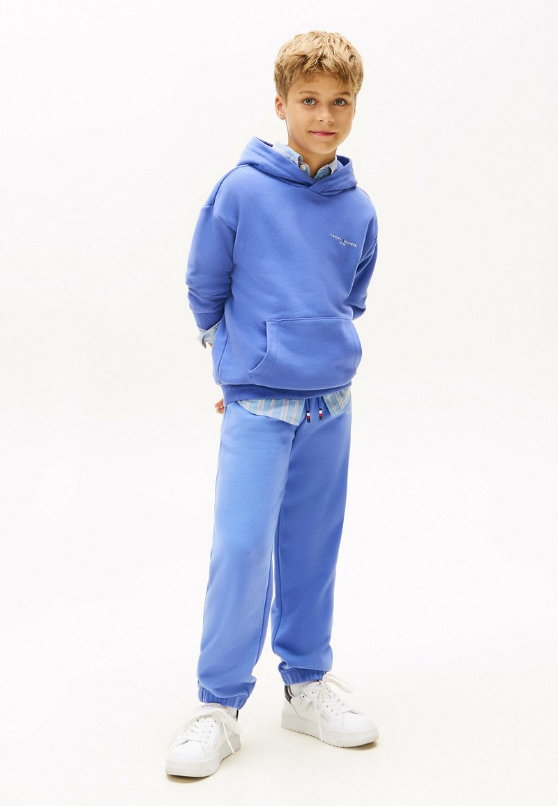 Blå sweatshirt med hætte, frontlomme og logo; matchende blå sweatpants med elastiske manchetter; hvide sneakers; lagdelt med skjorte med krave.