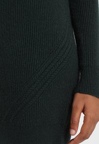 Jersey de punto de color verde oscuro con textura en diagonal y mangas ajustadas, que muestra una tela suave y cálida.