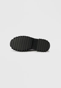 Black rubber shoe sole with a tread pattern, featuring ridges for traction and a rectangular branding label near the heel.