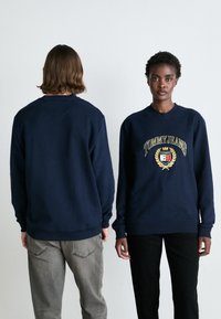 Sudadera de cuello redondo en color azul marino, confeccionada en algodón, con un emblema dorado que dice "TOMMY JEANS" en el frente, y con puños y dobladillo acanalados.