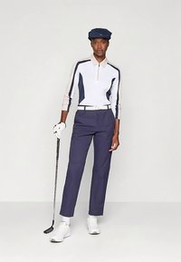 Γυναίκα γκολφ παίζει με στολή σε ναυτικό μπλε και άσπρο, κρατώντας τη λέιζα, φορώντας καπέλο, άσπρα γάντια, άσπρη ζώνη και άσπρα αθλητικά παπούτσια σε απλό φόντο.