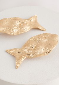Two gold fish-shaped ornaments with a hammered texture and varying surface patterns, resting on a light-colored pedestal.