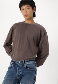 Camisa larga de manga larga marrón con un corte relajado, que presenta un cuello redondo. Combinada con unos vaqueros de mezclilla azules que tienen un lavado desgastado y un diseño de cinco bolsillos.