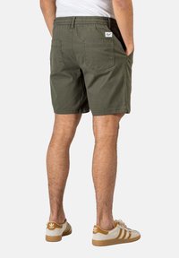 Pantalones cortos de algodón verde oliva con cinturilla elástica, dos bolsillos traseros y una etiqueta blanca; combinados con zapatillas en tonos crema y marrón.