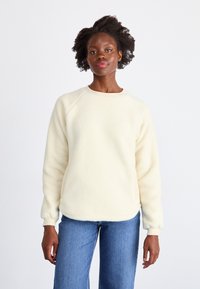 Felpa in pile crema con scollo rotondo, maniche raglan e dettagli a costine ai polsini, indossata con jeans in denim blu.