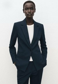 Marineblauwe blazer van gestructureerde stof, met een enkele knoopsluiting, ingezette revers en twee voorzakken, gecombineerd met een wit shirt eronder.