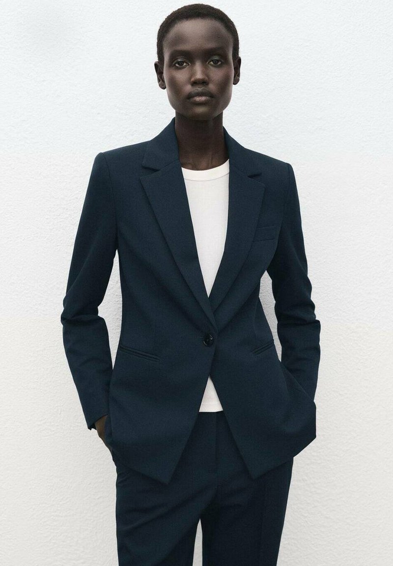 Marineblauwe blazer van gestructureerde stof, met een enkele knoopsluiting, ingezette revers en twee voorzakken, gecombineerd met een wit shirt eronder.