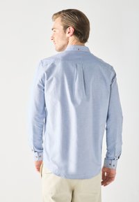 Light blue button-up shirt with long sleeves, featuring a pleated back, contrasting buttons at cuffs, and a curved hemline.