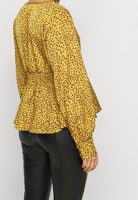 Gul blus med brunprickigt mönster; lång ärm, peplumslut, smockad midjedetalj, lättviktsmaterial och rund halsringning.