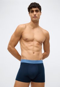 Muscular man with short curly hair wearing navy blue boxer briefs with a light blue waistband, standing against a plain light background.