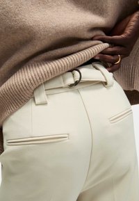 Beige gebreide trui die gedeeltelijk een hooggetailleerde crèmekleurige broek bedekt met een gladde textuur, inclusief een riem en achterzakdetails.