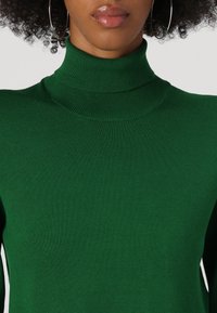 Pull en col montant côtelé vert fait de tissu tricoté extensible; présente un col haut et une silhouette ajustée.