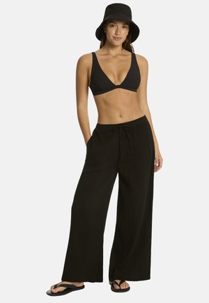 Kobieta w czarnym kapeluszu typu bucket, czarnym bikini topie, luźnych czarnych spodniach i czarnych klapkach stojąca na białym tle.