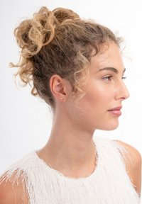 Krullend haar in een losse opgestoken stijl, met kleine oorringen en een steker. Draagt een textuurrijk wit mouwloos topje met rafelige randen.