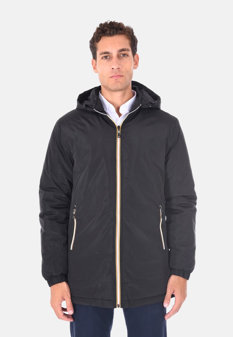 Uomo che indossa una giacca nera con cappuccio e dettagli in crema sulla cerniera, sopra una camicia bianca, in piedi contro uno sfondo chiaro e uniforme.