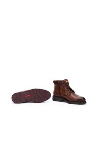 Botas de cuero marrón hasta el tobillo con un acabado texturizado, puntera redonda, cremallera lateral y un patrón de suela resistente. Incluyen un acento rojo en el talón.