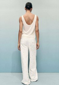 Top sans manches blanc avec un dos plongeant, associé à un pantalon blanc orné de petites broderies florales jaunes. Tissu lisse, coupe décontractée.