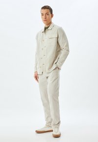 Lichtbeige linnen jas met grote voorknoop, een borstzak, gecombineerd met bijpassende broek en witte sneakers met bruine accenten.