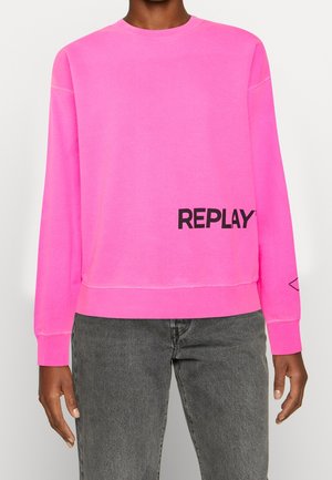 Sweat-shirt à manches longues rose vif avec le texte noir "REPLAY" en bas à droite sur le devant, porté avec un jean gris délavé.