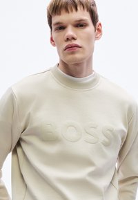 Μπεζ φούτερ με υφασμάτινο, τονικό λογότυπο "BOSS". Διαθέτει στρογγυλή λαιμόκοψη και ριμπ μανσέτες. Λείο ύφασμα, άνετη εφαρμογή.