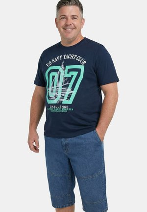 Uśmiechnięty mężczyzna w granatowym t-shircie z grafiką klubu jachtowego i dżinsowych spodenkach do kolan, stoi z jedną ręką w kieszeni.