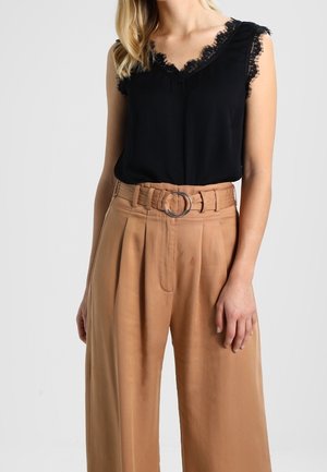 Femme portant un haut noir sans manches orné de dentelle, rentré dans un pantalon large taille haute couleur beige avec une boucle de ceinture circulaire.