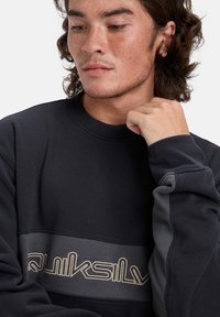 Czarna bluza z teksturowanym wykończeniem. Posiada poziomy szary pasek oraz kontrastowe beżowe logo "Quiksilver" na piersi.
