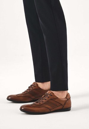 Ruskeat nahkaiset rennot nauhalliset kengät, joita käytetään mustien kapeiden housujen kanssa vaalealla taustalla.