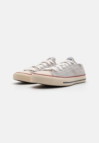Pantofi sport din pânză de culoare gri deschis, cu vârf rotund, talpă din cauciuc alb și dungi de accent roșii și bleumarin. Prezintă un design clasic cu șireturi.