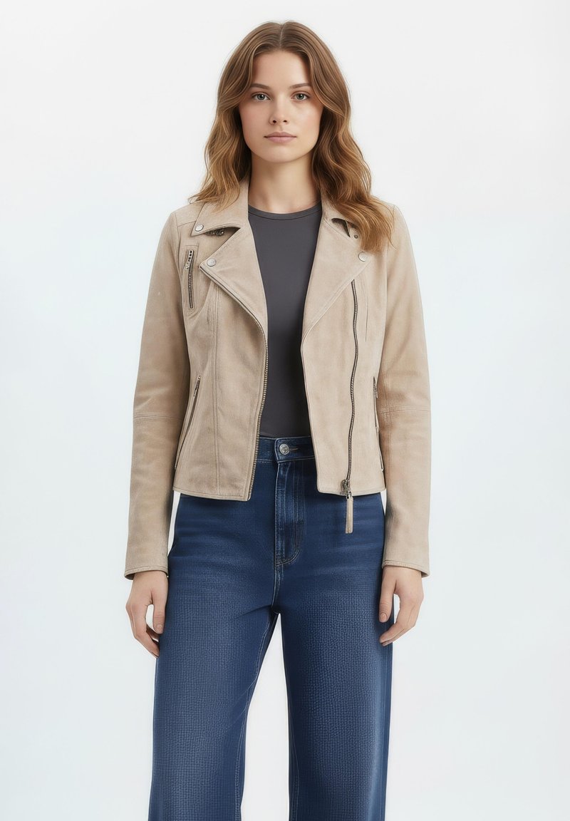 Vrouw met golvend lichtbruin haar, gekleed in een beige suède jas, een donkergrijze top en hooggetailleerde donkerblauwe spijkerbroek, staand tegen een witte achtergrond.