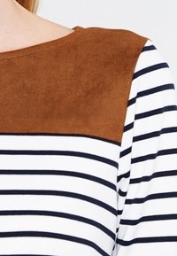 Randig långärmad topp med en brunt mockaliknande axelpanel, samt marinblå ränder på en vit bakgrund. Mjuk textur överlag.