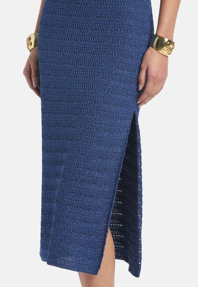 Jupe en maille texturée bleue avec fente latérale portée par une personne avec des bracelets manchettes dorés aux deux poignets sur fond blanc.