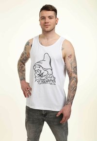 Canotta bianca con una grafica frontale di un gnome cartone animato etichettato "FELICE". Indossata con jeans grigi strappati; presenta tatuaggi visibili sulle braccia.