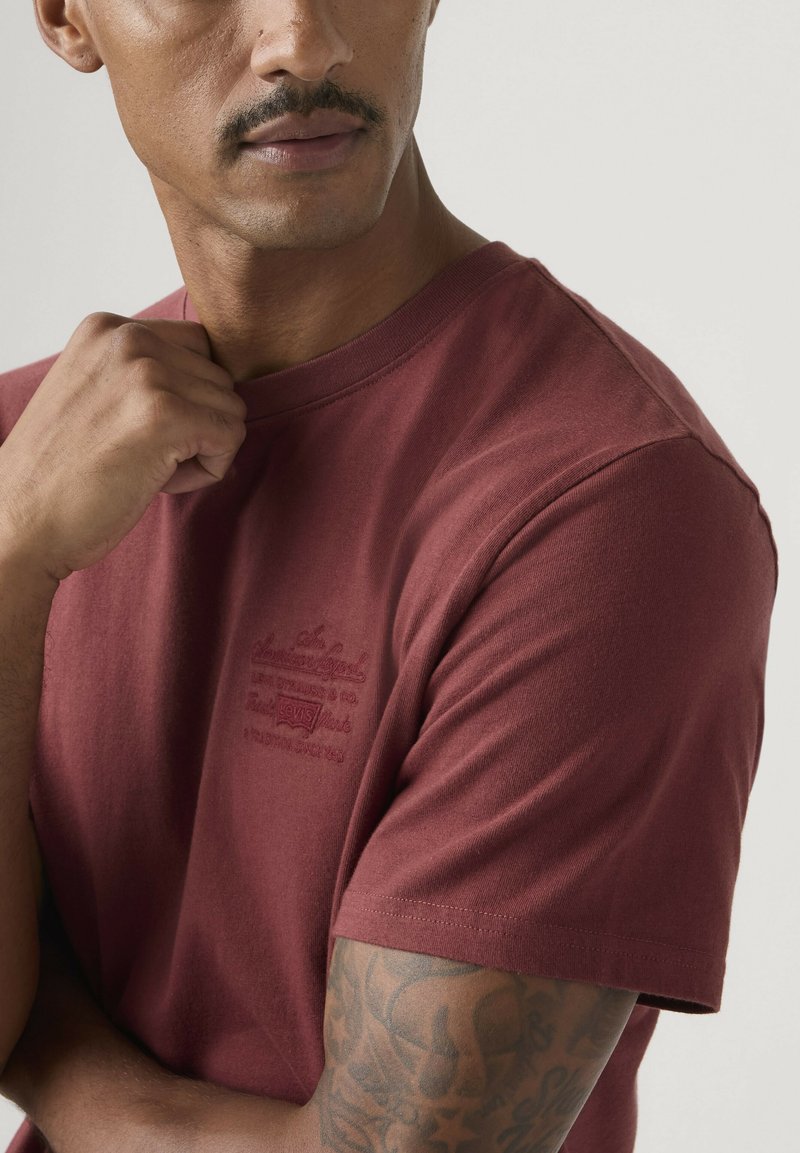 Mand med tatoveret arm iført en vinrød kortærmet T-shirt, der holder kraven med sin højre hånd mod en neutral baggrund.