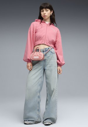 Mujer joven con sudadera rosa corta con capucha, pantalones anchos de mezclilla azul claro, zapatos plateados y un pequeño bolso rosa de Puma colgado del lazo del cinturón.