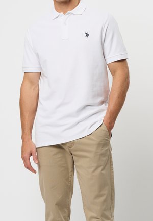 Homme portant un polo blanc avec un petit logo sombre sur la poitrine et un pantalon beige, debout avec une main dans la poche.