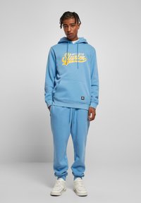 Felpa leggera blu con testo grafico giallo, dotata di tasca frontale e lacci. Abbinata a pantaloni joggers blu chiaro e sneakers bianche.