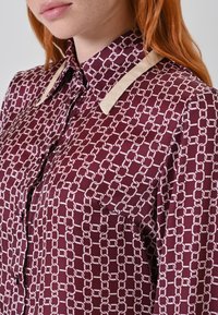 Blusa bordeaux con stampa a catena, caratterizzata da una texture in satin lucido, accenti color crema sul colletto e dettagli con bottoni lungo la parte anteriore.