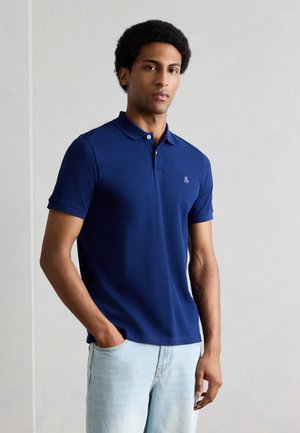 Άνδρας με κοντά σγουρά μαλλιά, φορώντας μπλε ναυτικό πόλο πουκάμισο και ανοιχτόχρωμα μπλε τζιν, στέκεται μπροστά σε ένα απλό φωτεινό τοίχο.