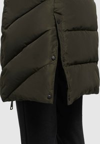 Donkergroene gewatteerde jas met drukknoopopening aan de zijkant, gedragen over zwarte broek, waarbij de voorkant van de outfit weergegeven wordt.