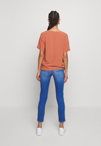 Top corto de color naranja quemado combinado con jeans ajustados azules y zapatillas blancas, visto desde atrás. La tela parece suave con un ajuste relajado.