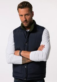 Marineblaue Steppweste mit grünem Fleecekragen, ausgestattet mit Druckknöpfen, einem gesteppten Design und einer glatten Textur, getragen über einem weißen Langarmshirt.