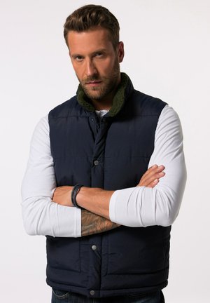 Marineblaue Steppweste mit grünem Fleecekragen, ausgestattet mit Druckknöpfen, einem gesteppten Design und einer glatten Textur, getragen über einem weißen Langarmshirt.
