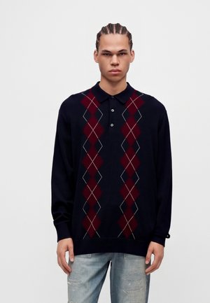 Jeune homme aux cheveux tressés portant un polo bleu marine à manches longues avec des motifs losanges rouges et un jean bleu clair déchiré.
