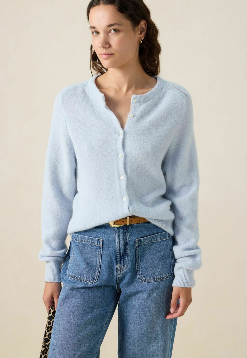 Femme portant un cardigan bleu clair à boutons avec un col festonné, un jean bleu avec de grandes poches avant, et tenant un sac à imprimé léopard.