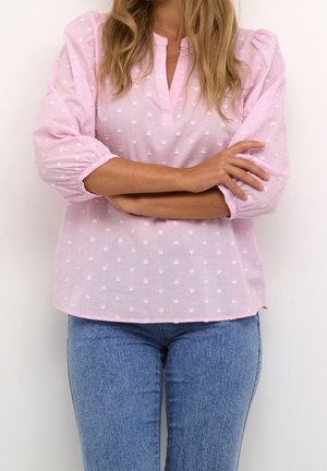 Woman with long hair wearing a light pink textured blouse with three-quarter sleeves and blue jeans, arms crossed at waist level.